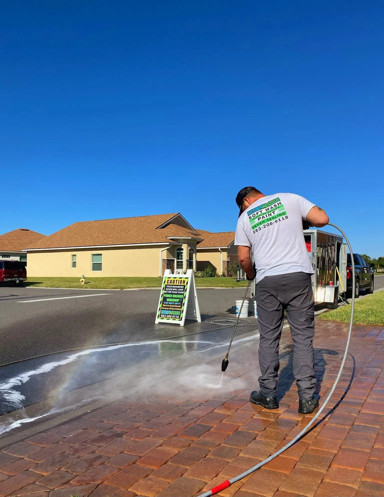 Worker pressure-washing a brick driveway beside a house under a clear blue sky
