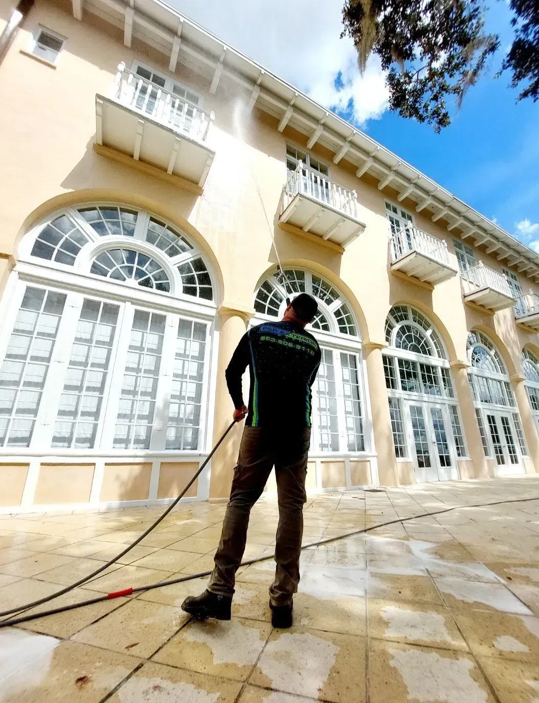 Person pressure-washing a cream-colored building with large arched windows and balconies under a blue sky