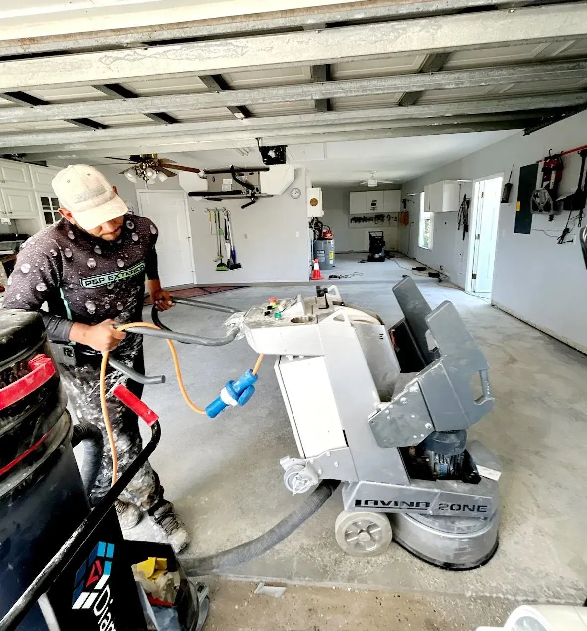 Worker using a floor grinder in a garage with exposed ceiling beams and tools nearby