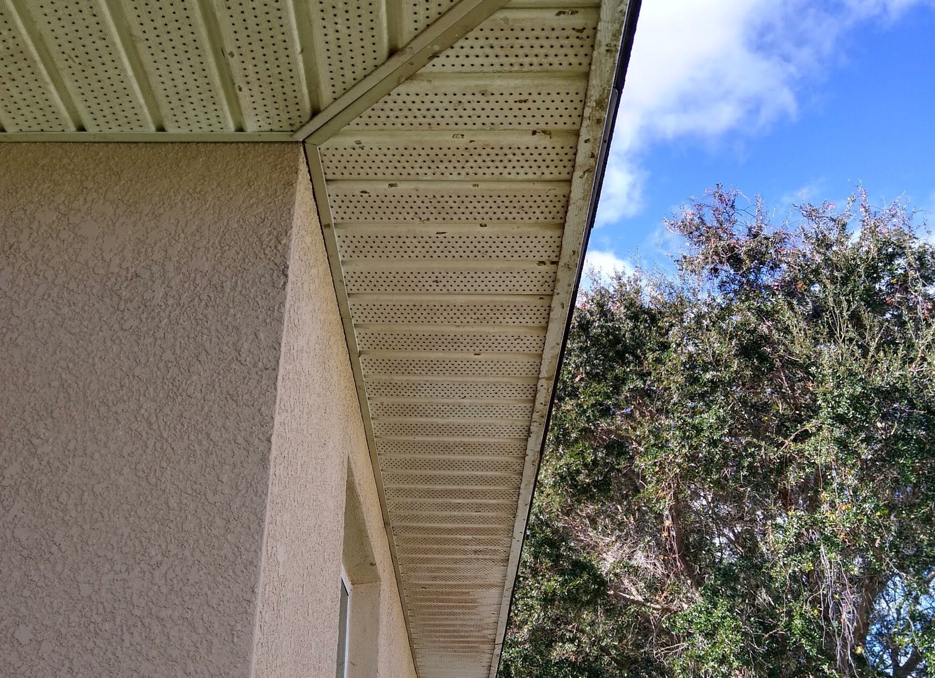 Corner of a beige stucco house with perforated eaves beside a tree and blue sky