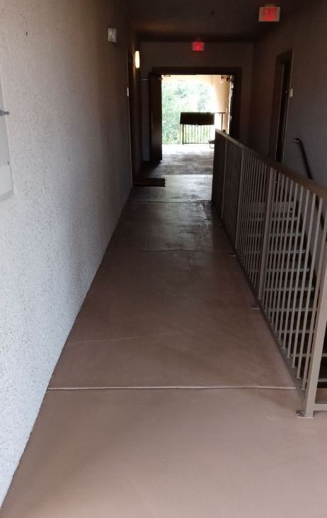 Narrow indoor corridor with beige floor, gray walls, and a metal railing leading to a bright exit.