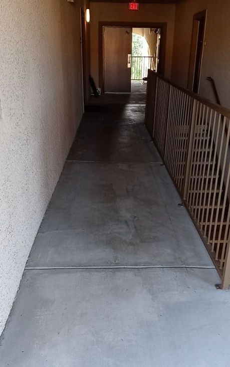 Empty apartment walkway with concrete floor, brown railing, and doorway at the end.