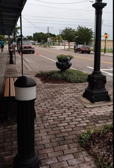 Brick sidewalk beside a quiet street with lampposts, shrubs, and parked cars under an overcast sky