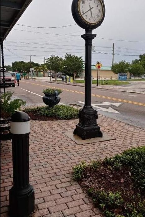 Brick sidewalk beside a black street clock and bollards, with a road and cloudy sky in the background.