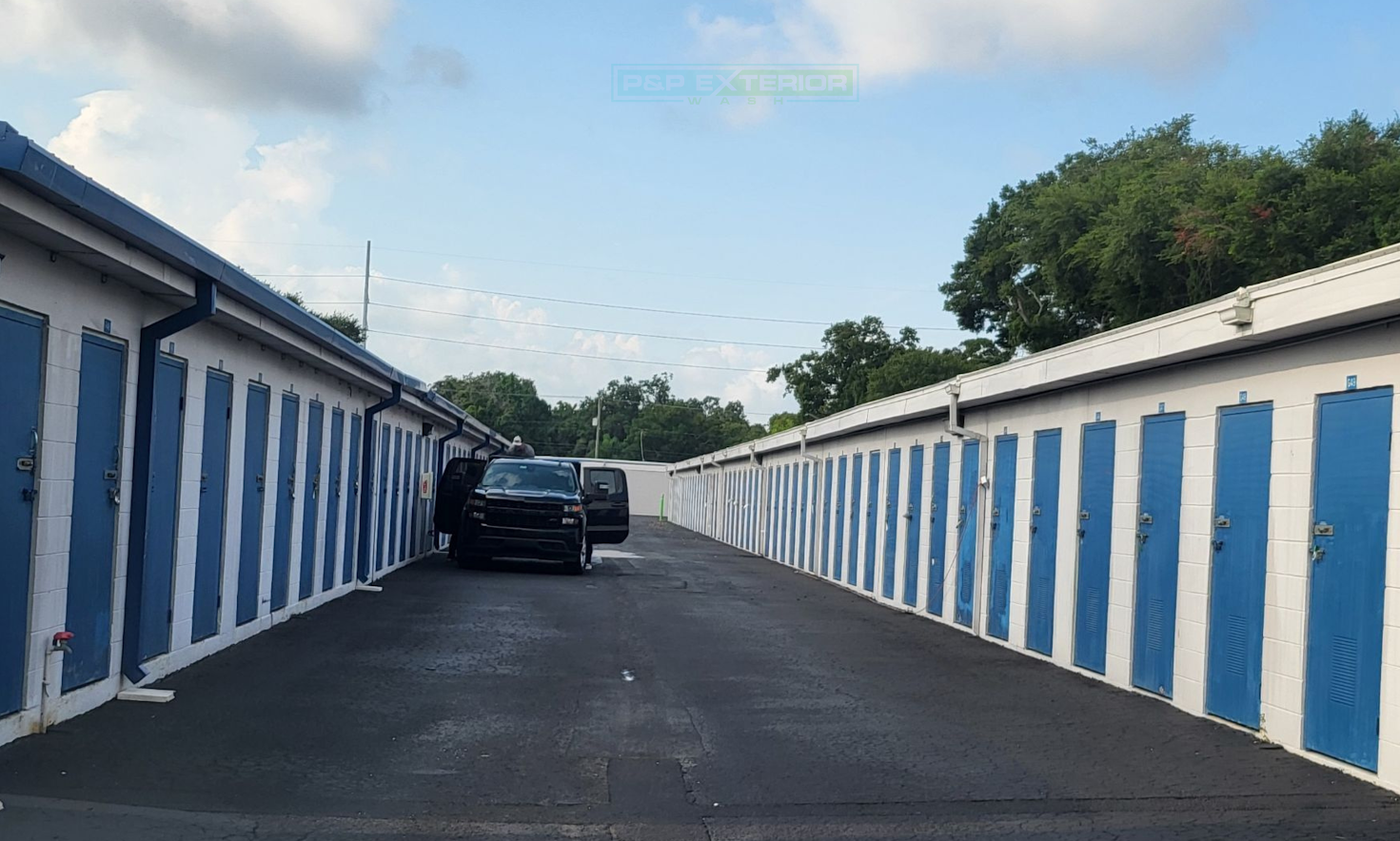 Row of blue-and-white storage units lining a driveway with a black SUV parked ahead under a cloudy sky
