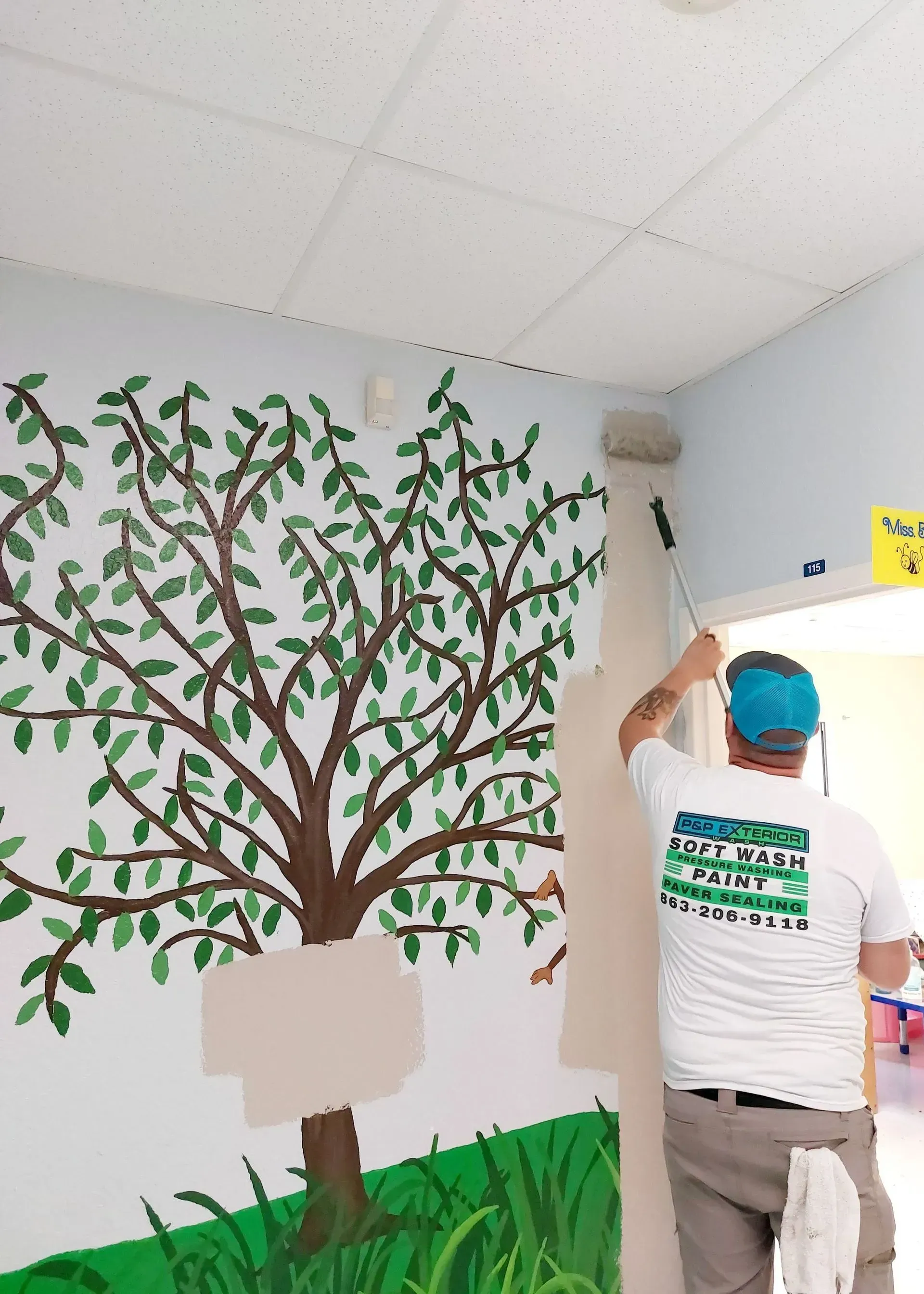 Person painting a classroom wall mural of a tree and grass, with a ladder nearby.