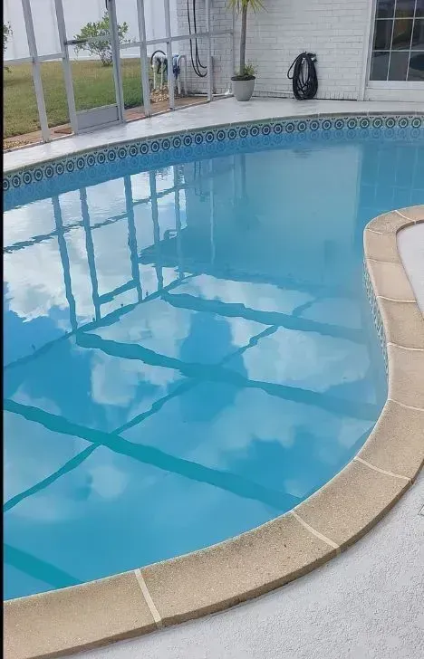 Curved blue swimming pool with beige coping and screened enclosure, reflecting windows and outdoor furniture.