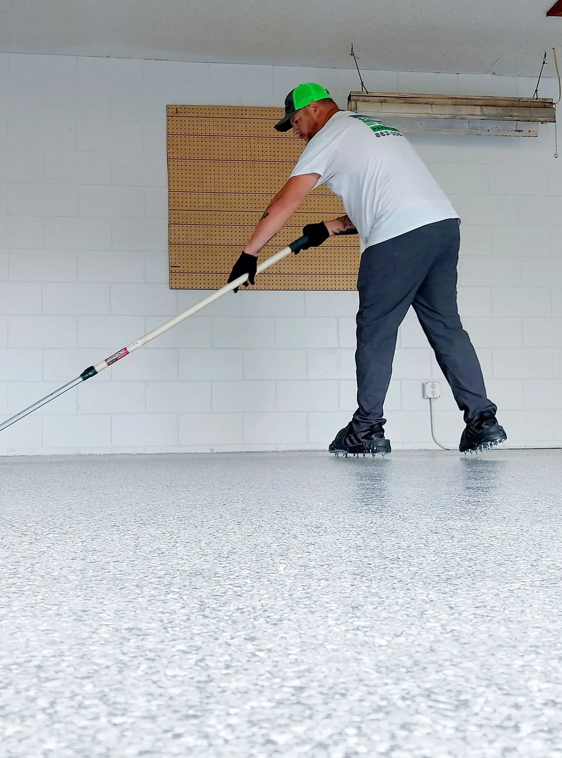 Worker smoothing a white floor with a long-handled tool in a room with cinderblock walls