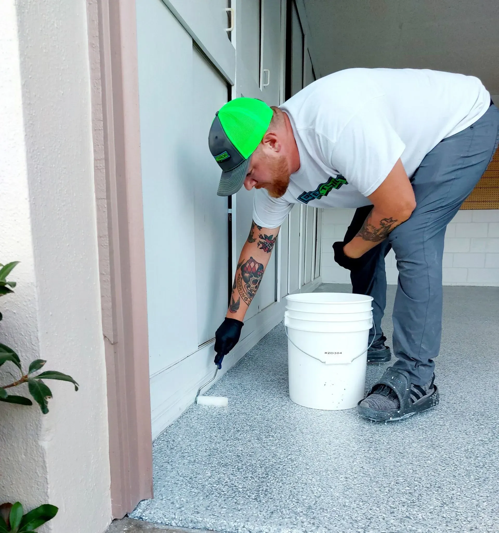 Person painting a doorway baseboard beside a white bucket, wearing a neon green cap and gloves.