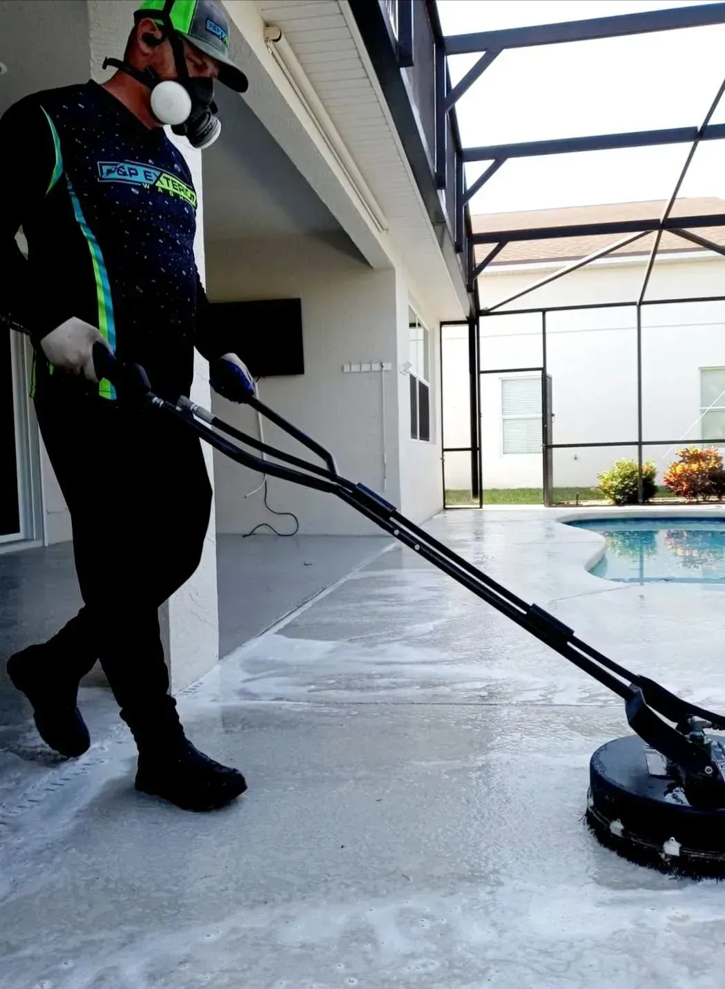 Person pressure-washing a patio beside a pool under a screened enclosure
