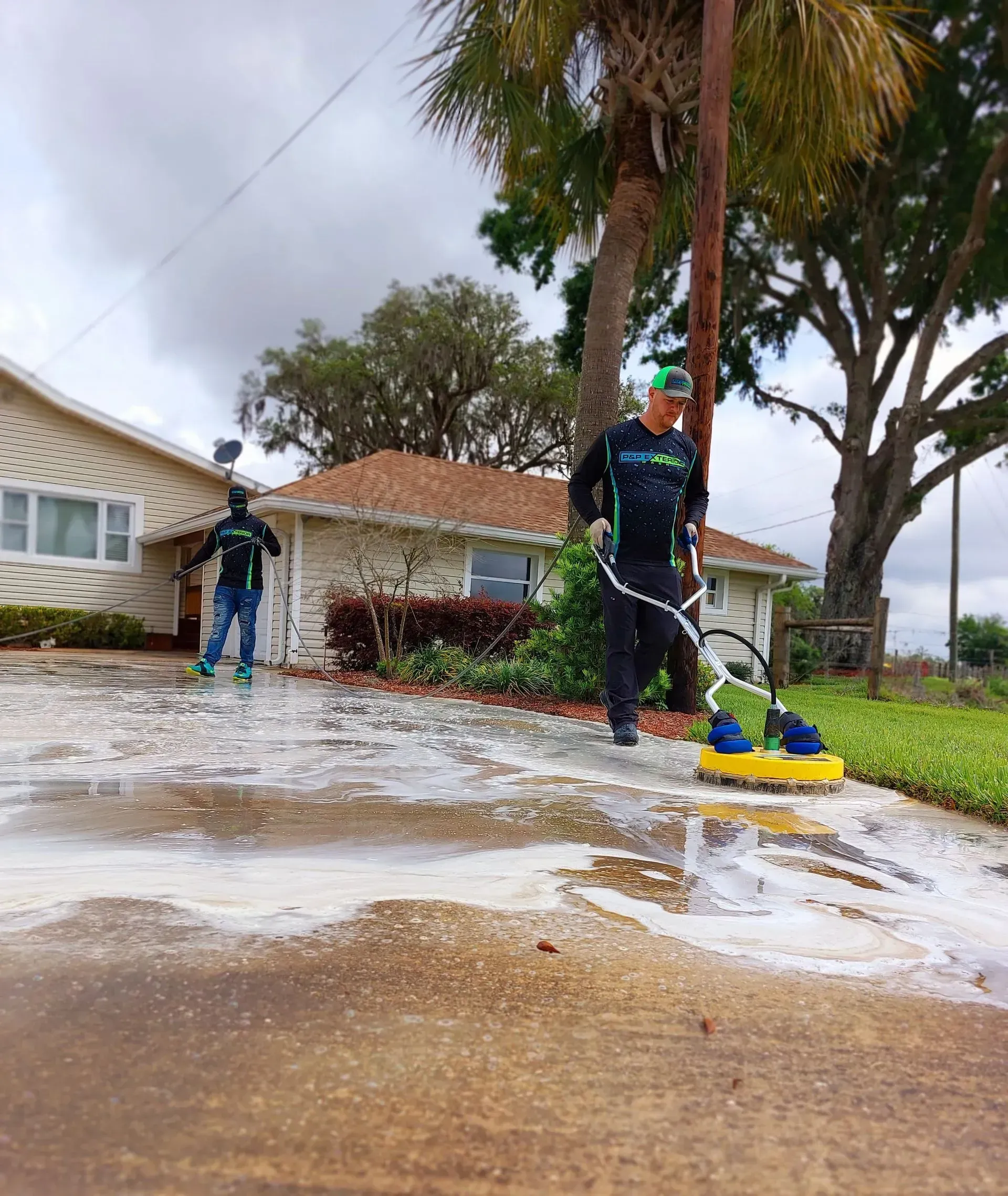 Workers pressure-washing a driveway outside a suburban house, with water spraying across the pavement.