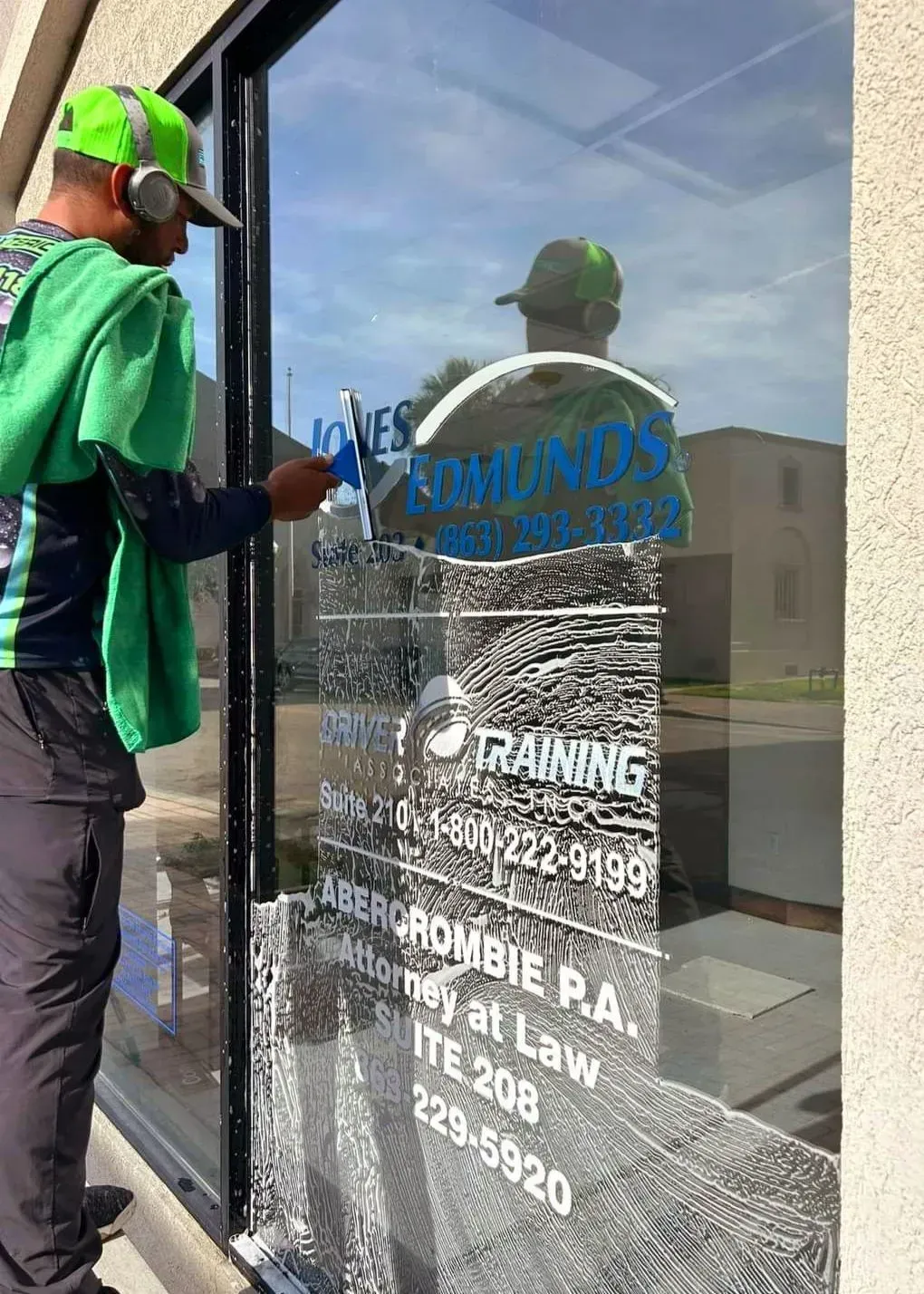 Worker in green shirt cleaning a glass door with business signs and a reflection of a building.