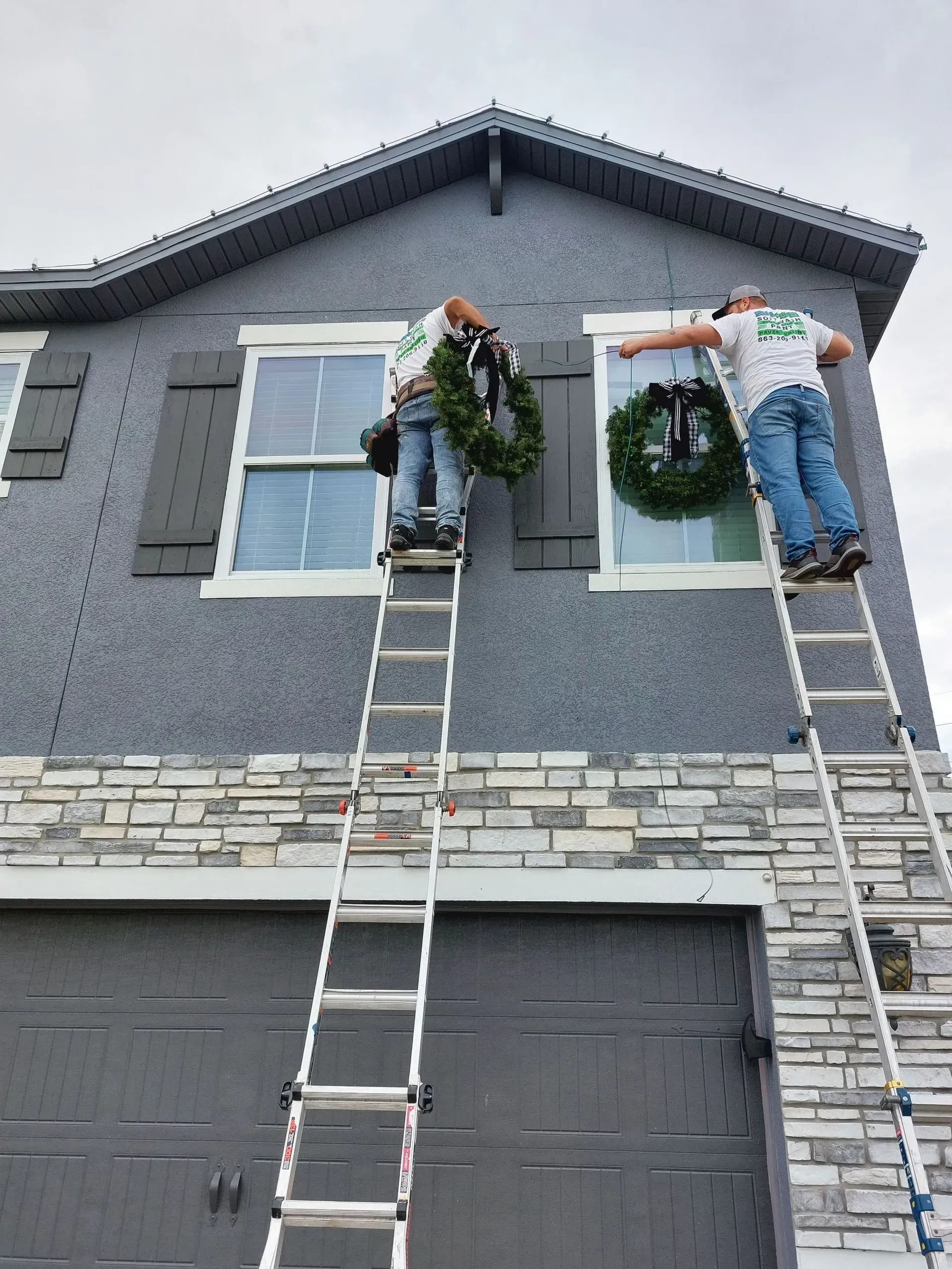 Two workers on ladders hang wreaths on a gray house with holiday garland.