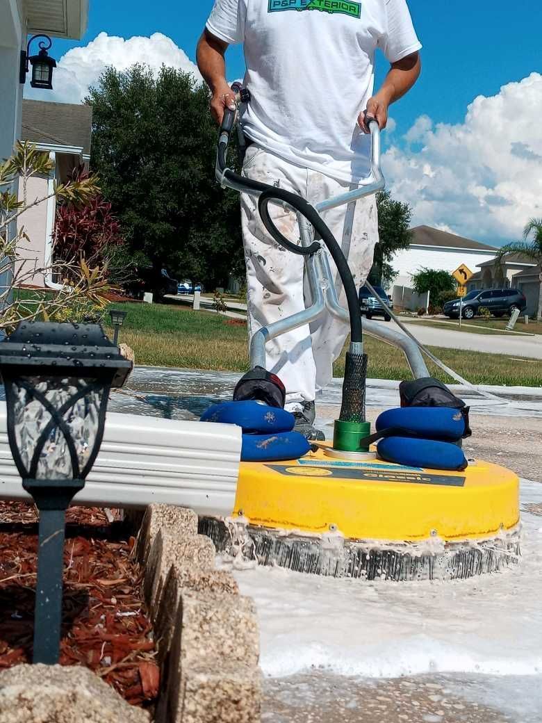 Worker pressure-washes a concrete surface with a yellow rotary cleaner on a sunny sidewalk.