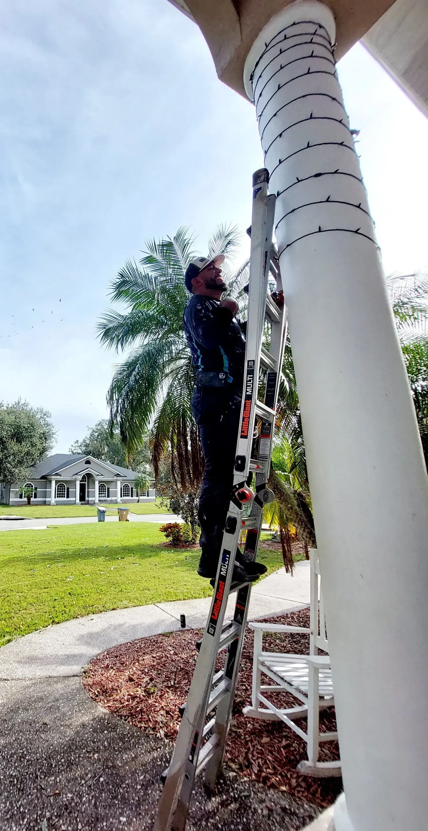 Worker on a ladder decorating a white porch column with string lights.
