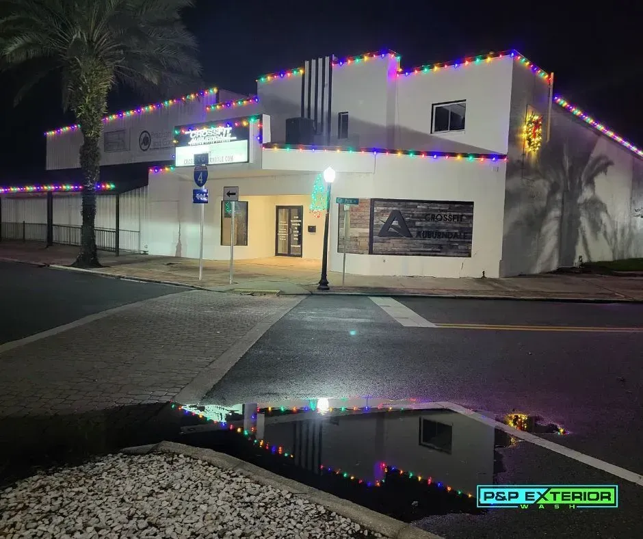 Nighttime view of a lit white commercial building with colorful roof lights reflected in a wet parking lot.