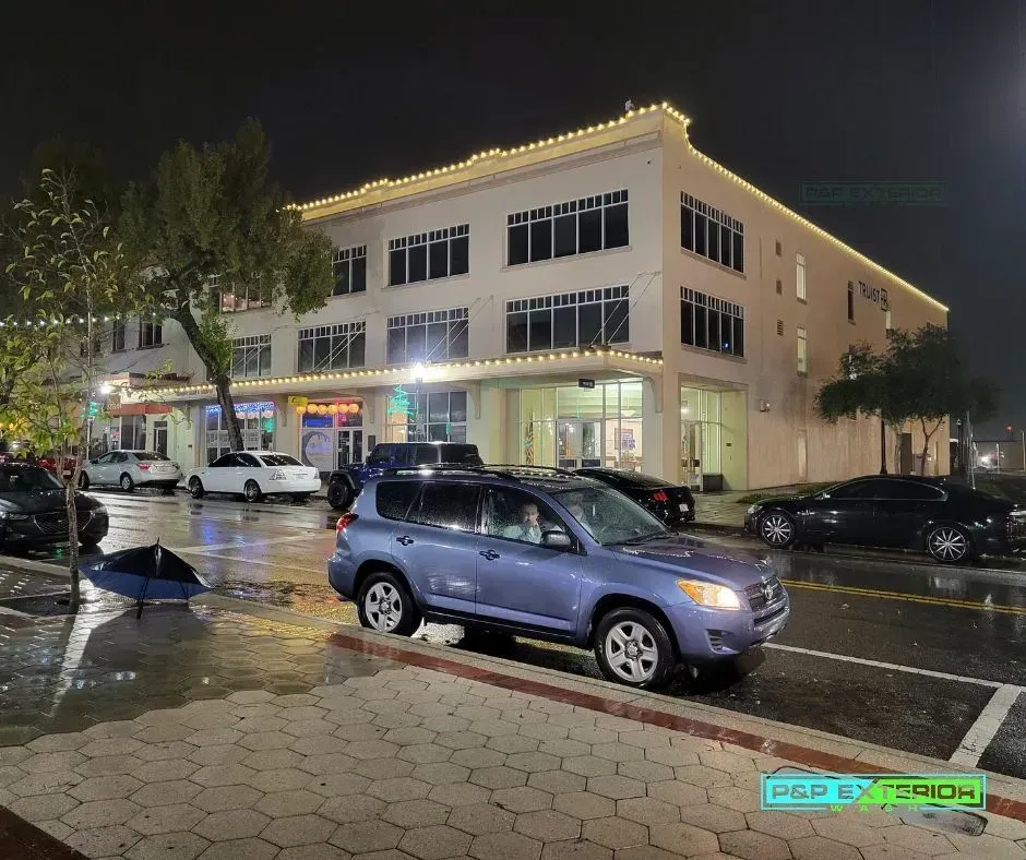 Nighttime street scene with a lit beige corner building and parked cars on a wet road.
