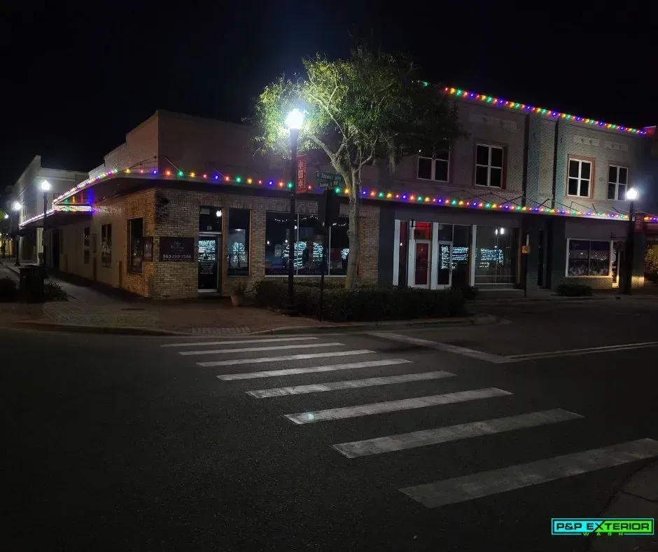 Nighttime street corner with a lit storefront, holiday lights, and a crosswalk in front.