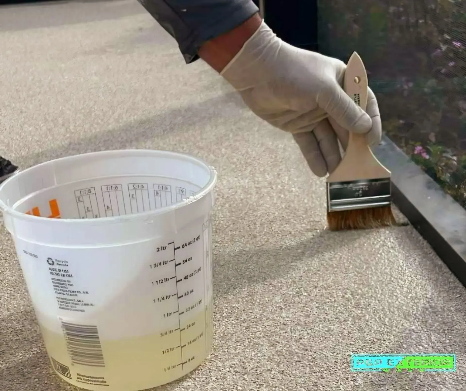 Gloved hand applying sealant with a brush beside a bucket on a concrete floor