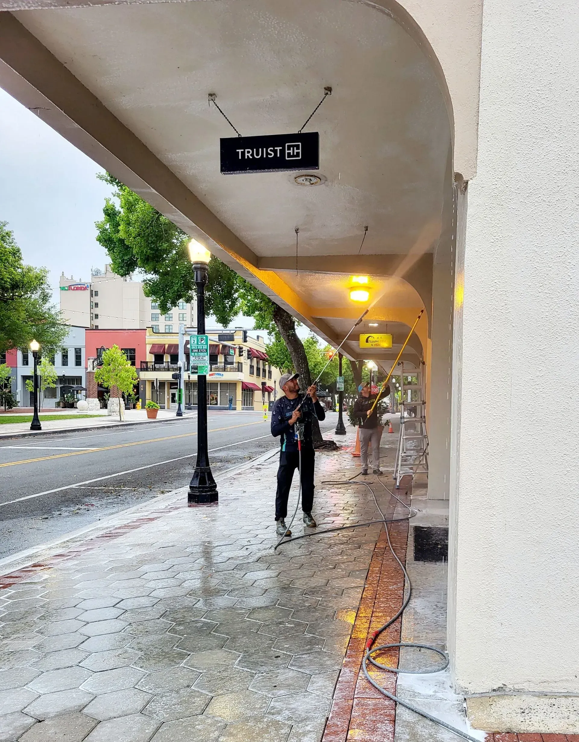 Rainy sidewalk under a covered arcade with two pedestrians and lit lamps along the street