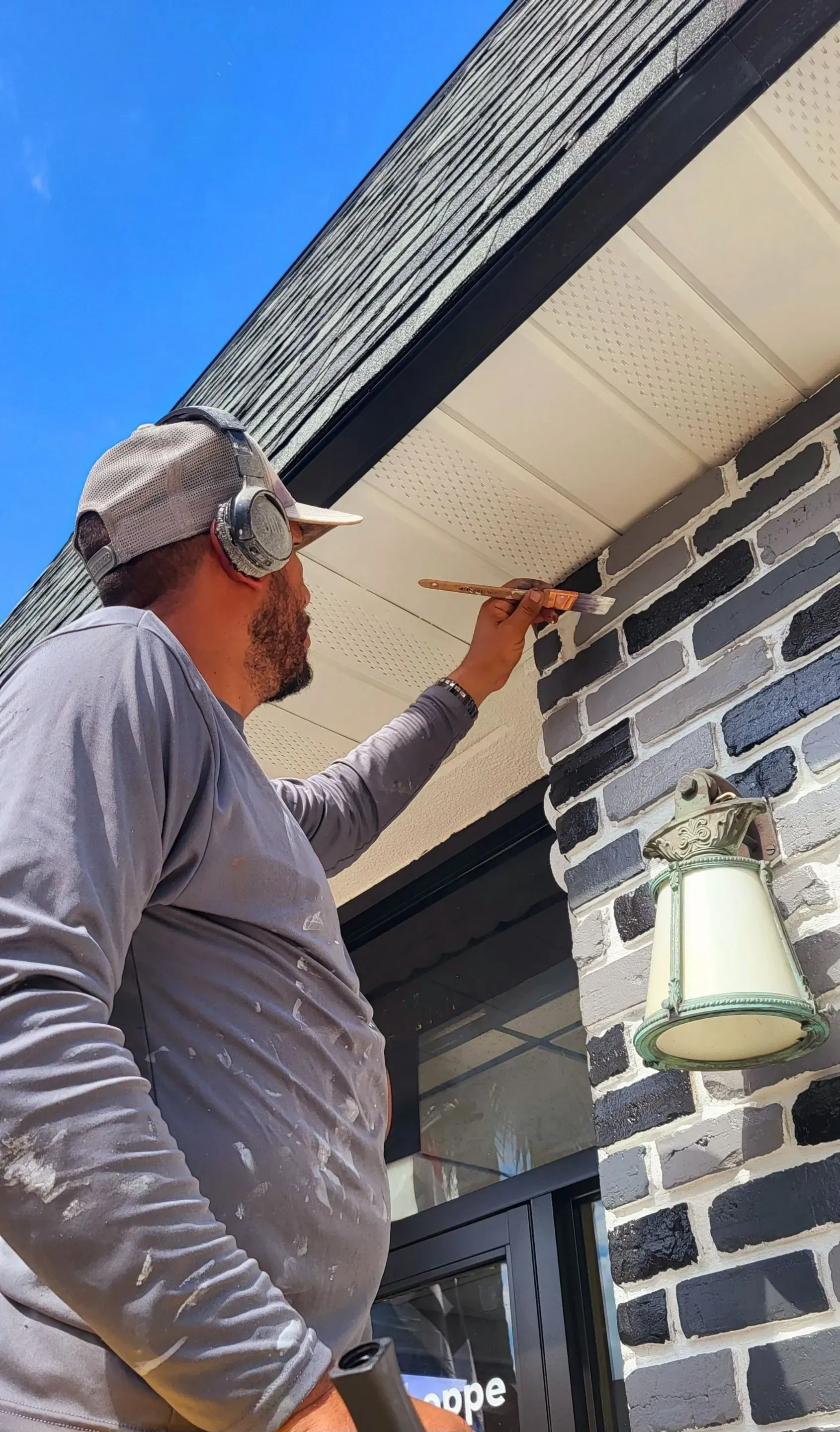 Person painting the underside of a house roof with a brush, standing on a porch ladder under a blue sky.