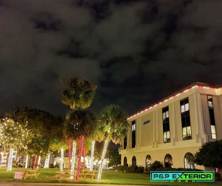 Nighttime exterior of a lit building with palm trees and red-white holiday lights on the lawn.
