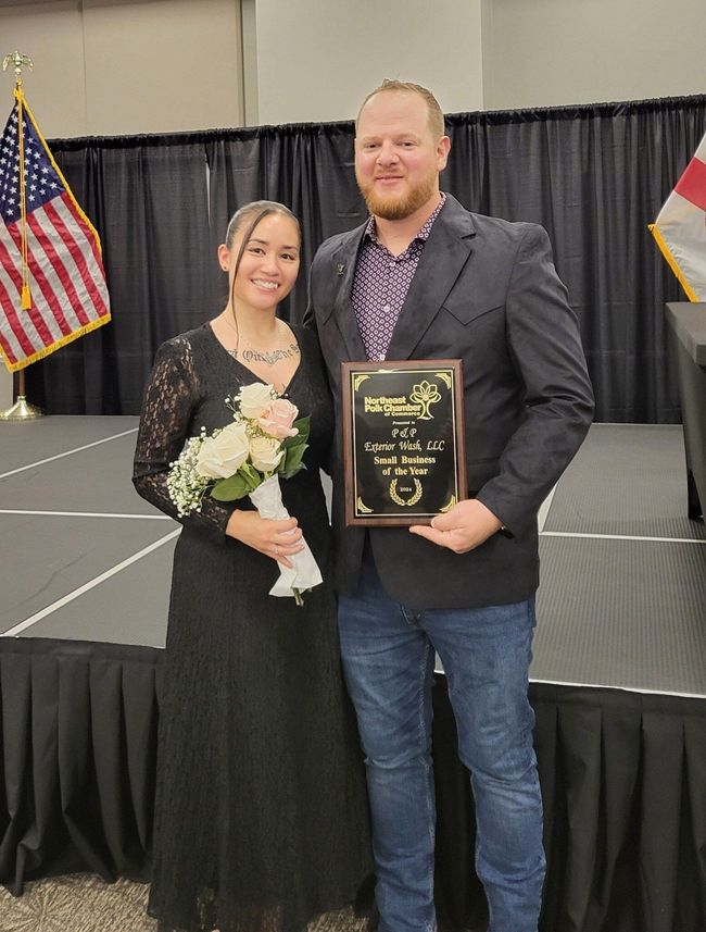 Two people on a stage holding a bouquet and plaque, with U.S. and state flags in the background.