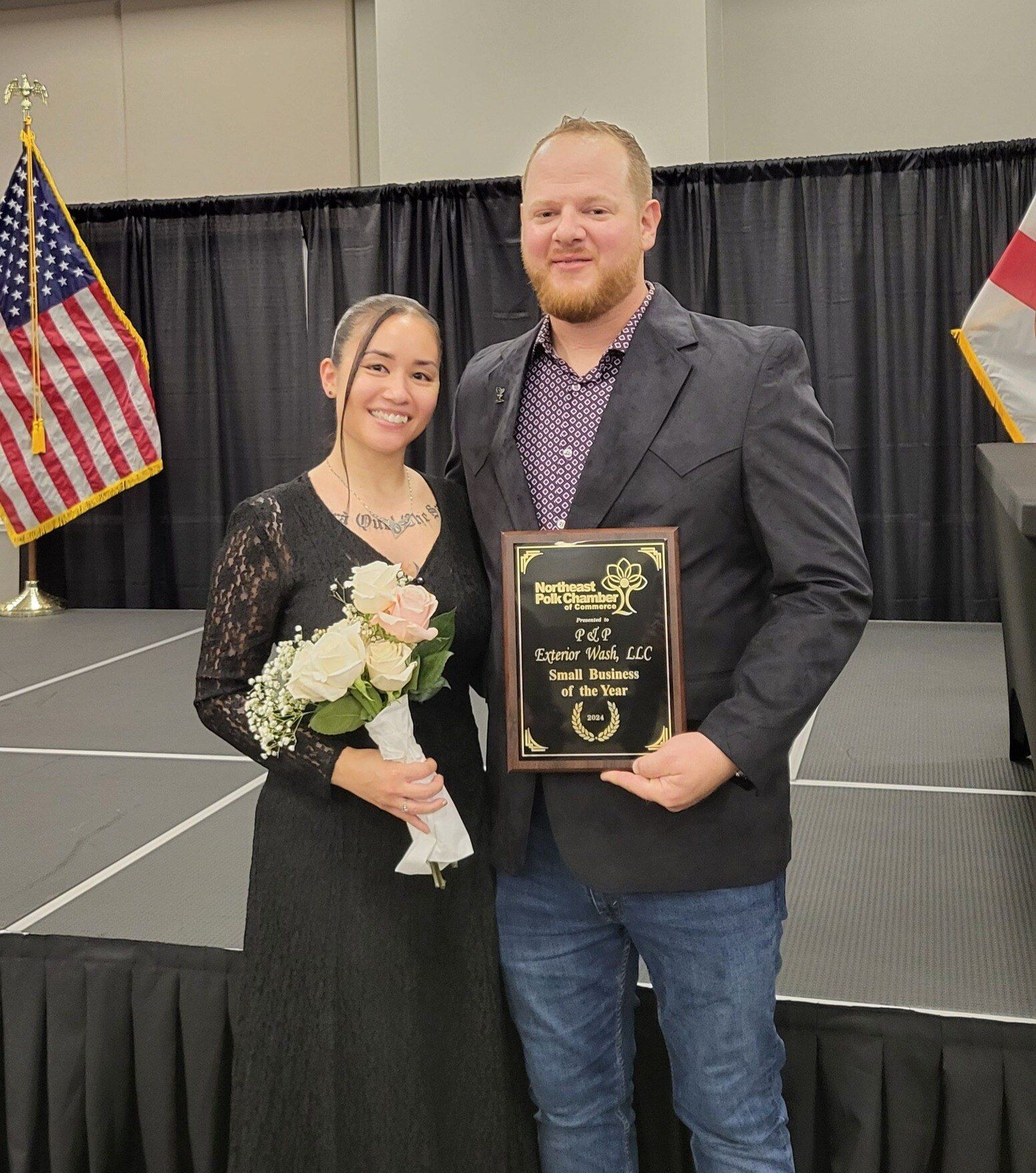 Two people posing onstage, holding a bouquet and plaque, with U.S. and flag backdrops.