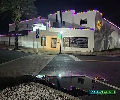 Nighttime view of a white two-story building outlined with colorful string lights, reflected in wet pavement.