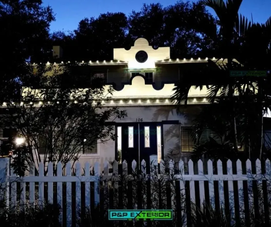 Nighttime view of a lit house behind a white picket fence and trees