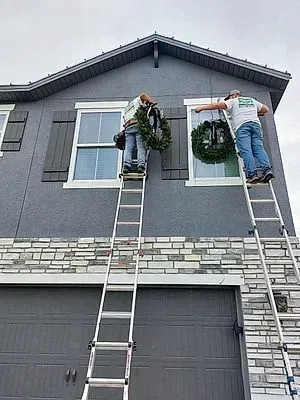 Two workers on ladders installing holiday wreaths on a gray house facade