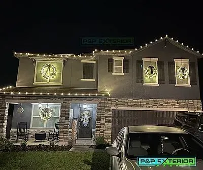 Two-story house at night, outlined in white Christmas lights with lit holiday wreaths and a parked car in front