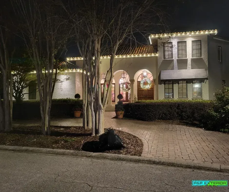 Night view of a lit two-story house with arched entry, brick driveway, and leafless trees.