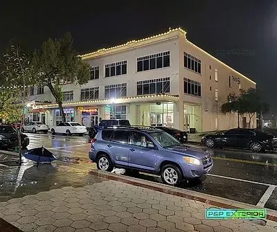 Nighttime street scene with a blue SUV parked in front of a lit two-story building after rain.