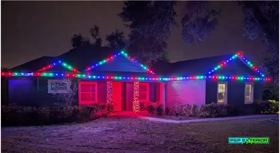 House at night outlined with colorful Christmas lights and red-lit windows, with a snowy yard.