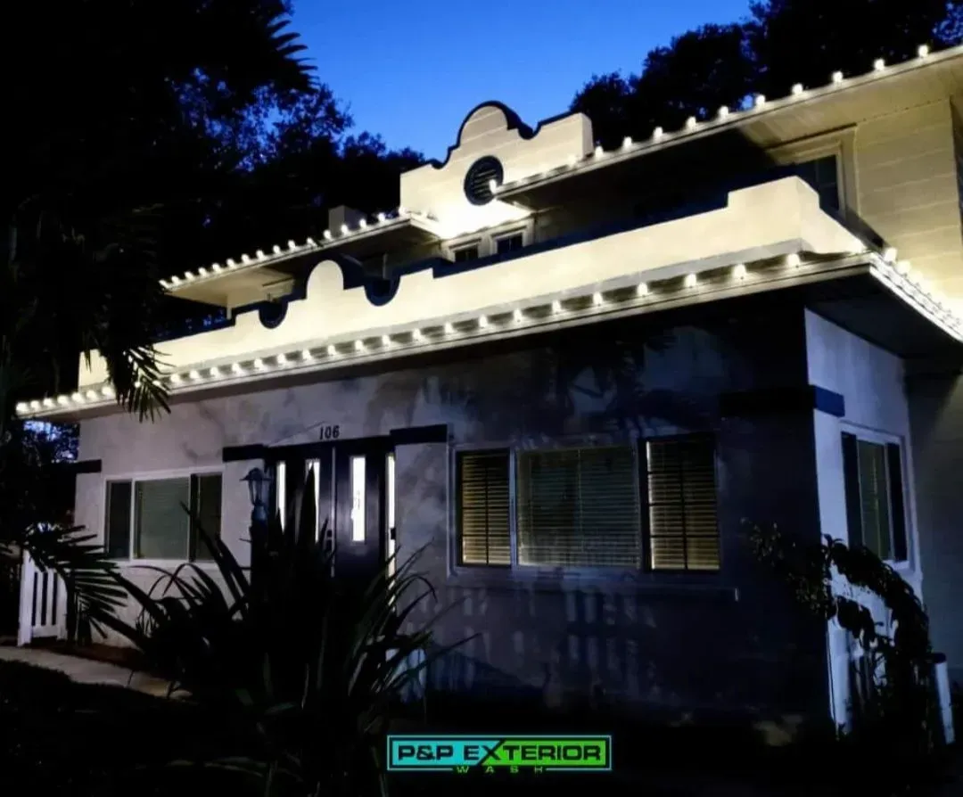 Nighttime view of a white house with lit facade, windows, and palm plants in front.