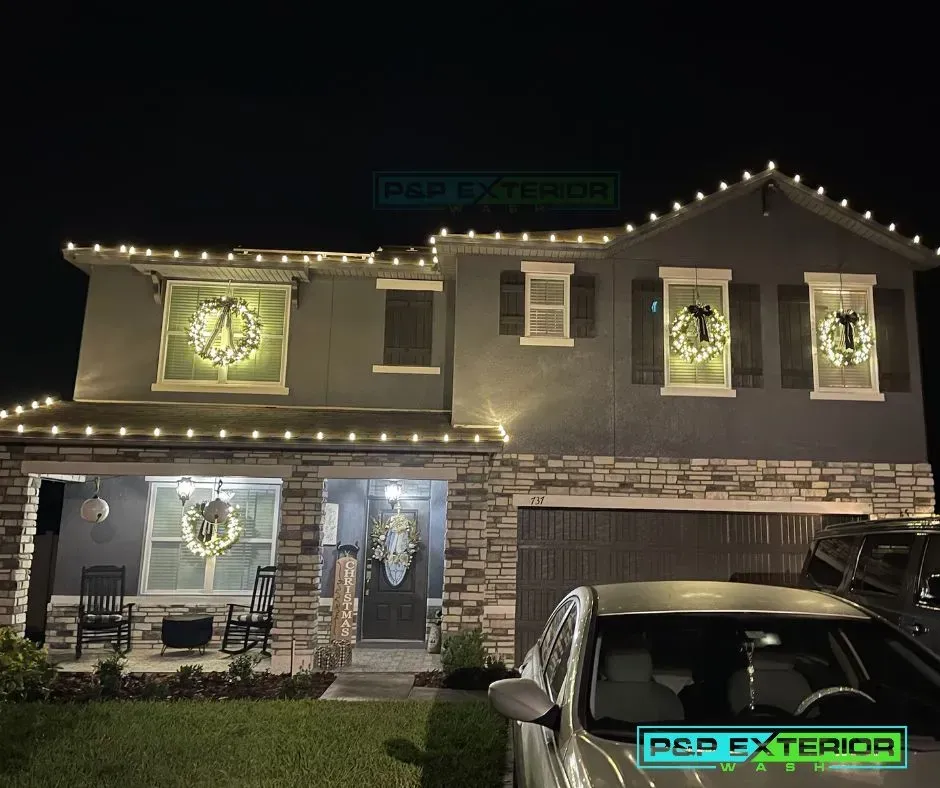 Two-story house at night outlined with string lights and festive window decorations, with cars parked in front.