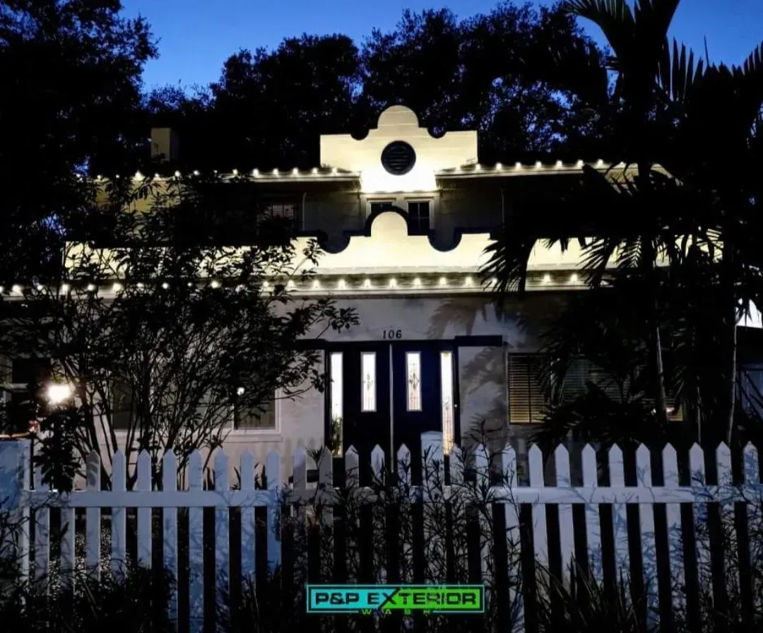 A white house at night, lit from within, behind a white picket fence and trees.