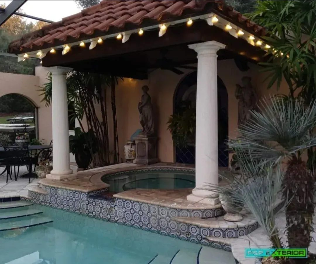 Tile fountain under a tiled patio with white columns, warm string lights, and tropical plants beside a pool