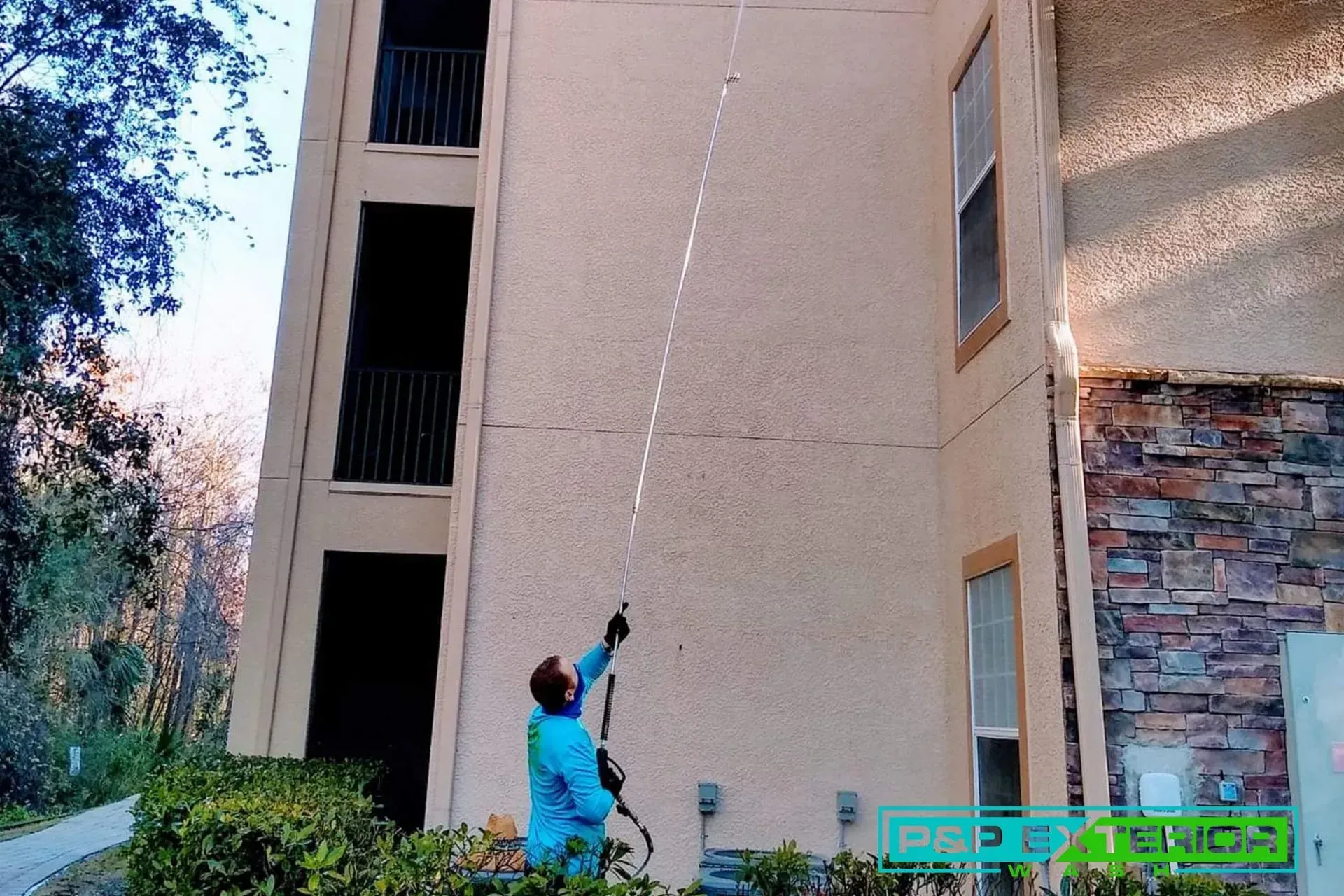 Person in blue spraying water up a tall apartment wall with a hose outdoors.