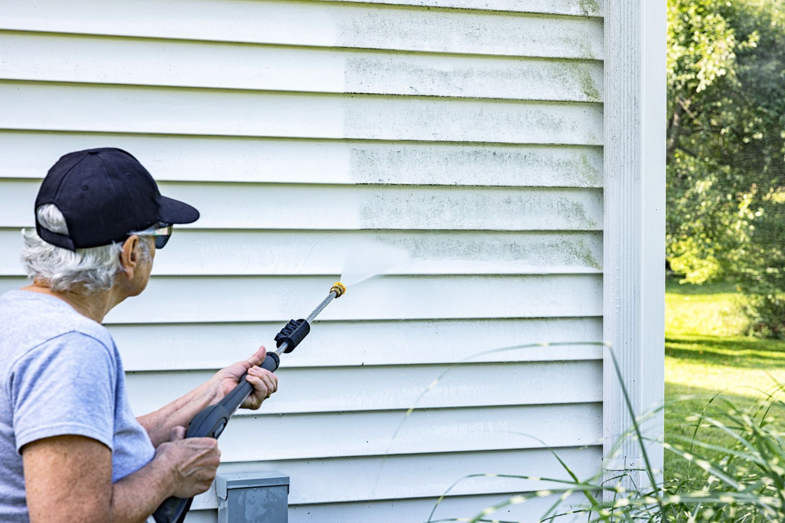 Person pressure-washing white siding beside a house, wearing a black cap and gray shirt