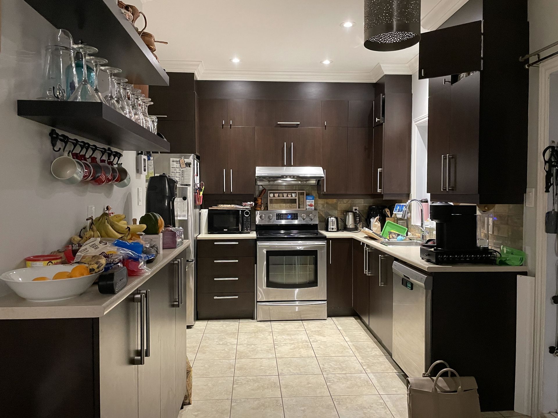 Cuisine en bois foncé avec appareils électroménagers en acier inoxydable, étagères avec verres et tasses et comptoir de couleur claire.