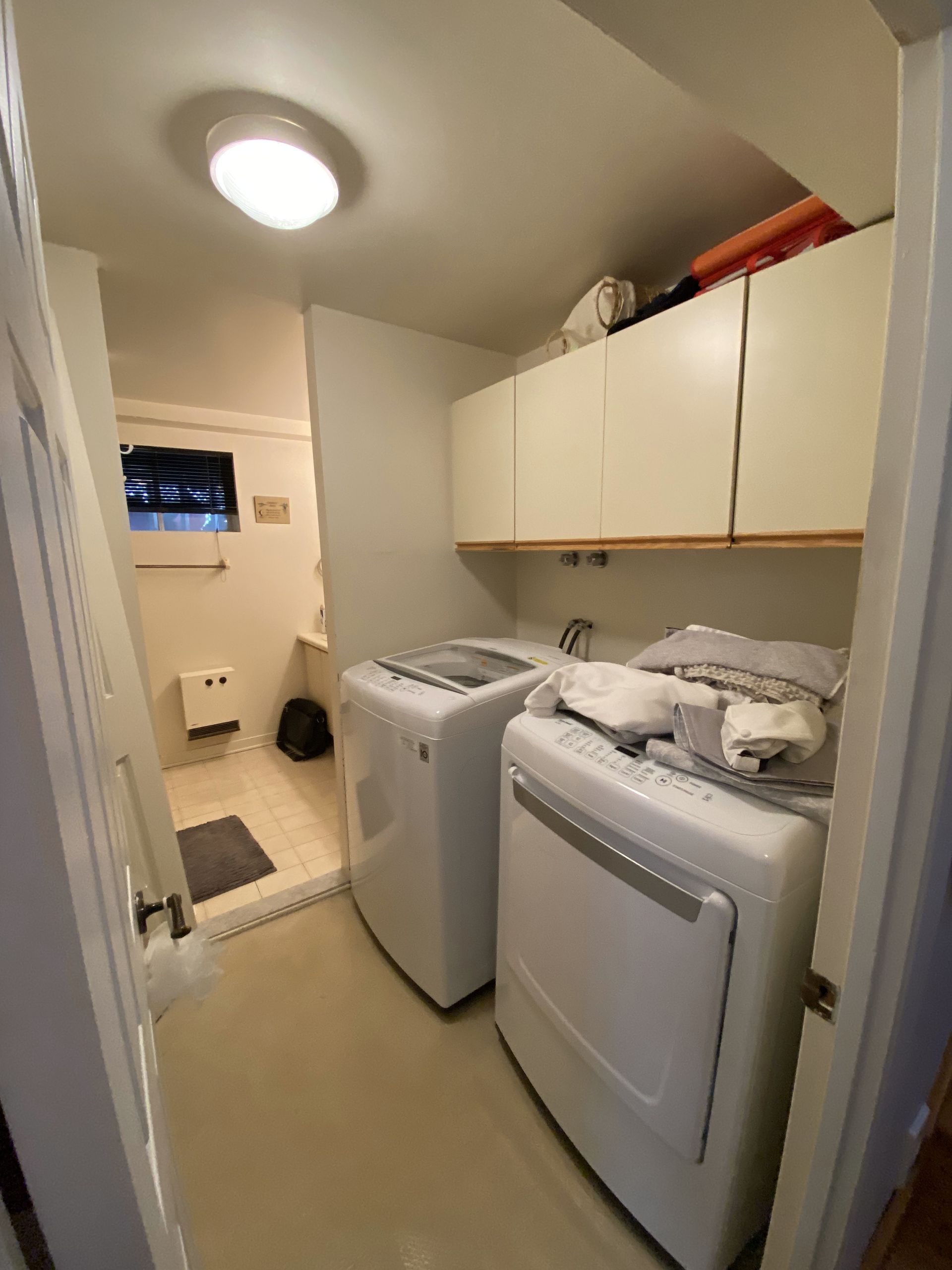 Buanderie avec laveuse et sécheuse blanches, armoires crème et une porte donnant sur une petite salle de bain.