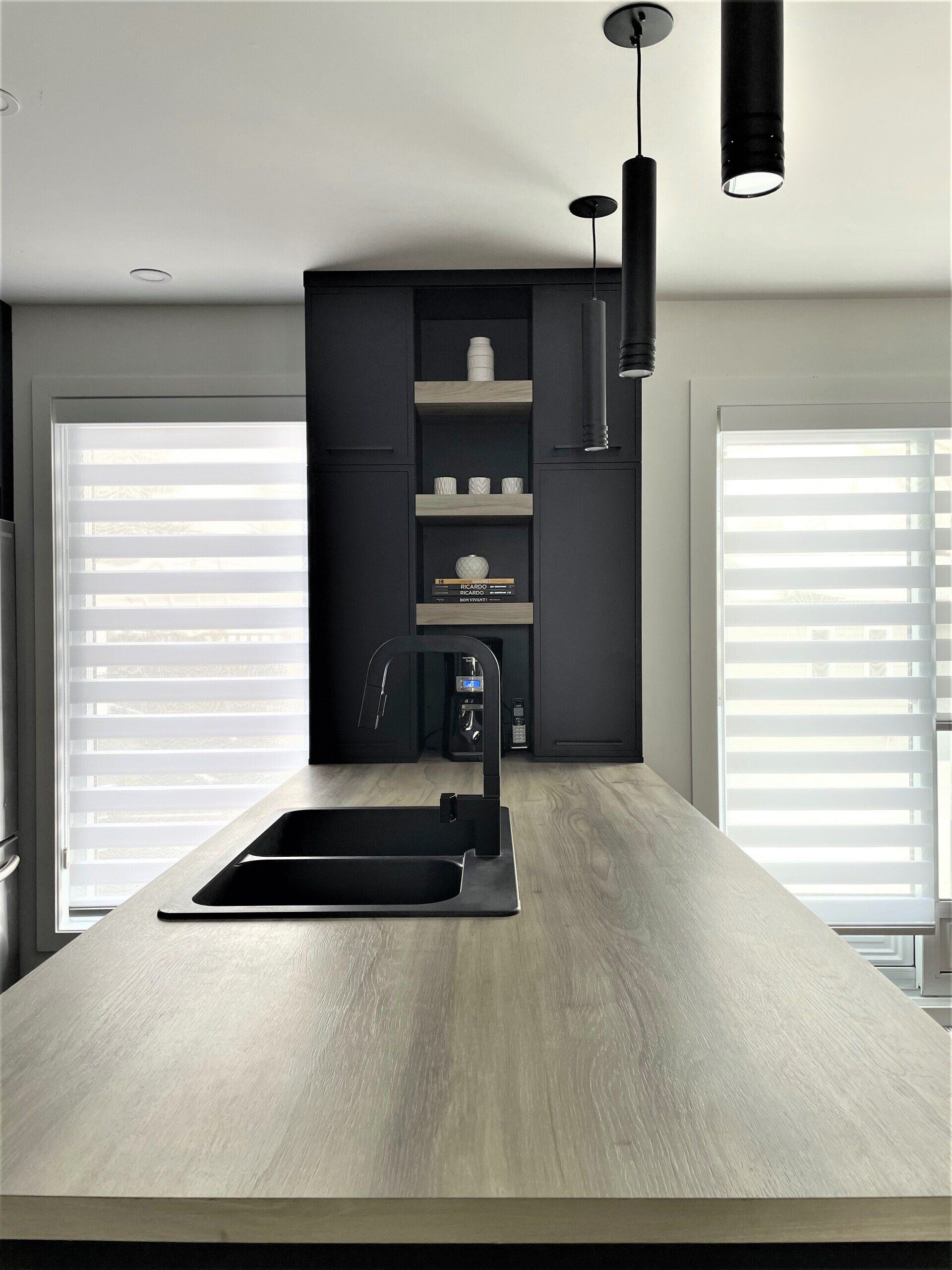 Cuisine avec évier et robinet noirs, îlot avec comptoir gris, armoires foncées et luminaires suspendus.