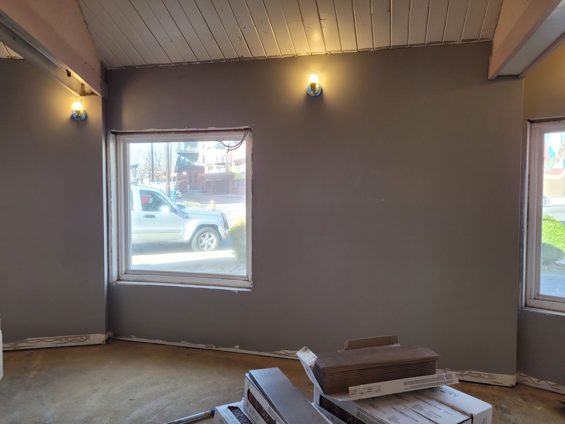 Empty room with gray walls, two windows, wall sconces, and a wooden floor under renovation.