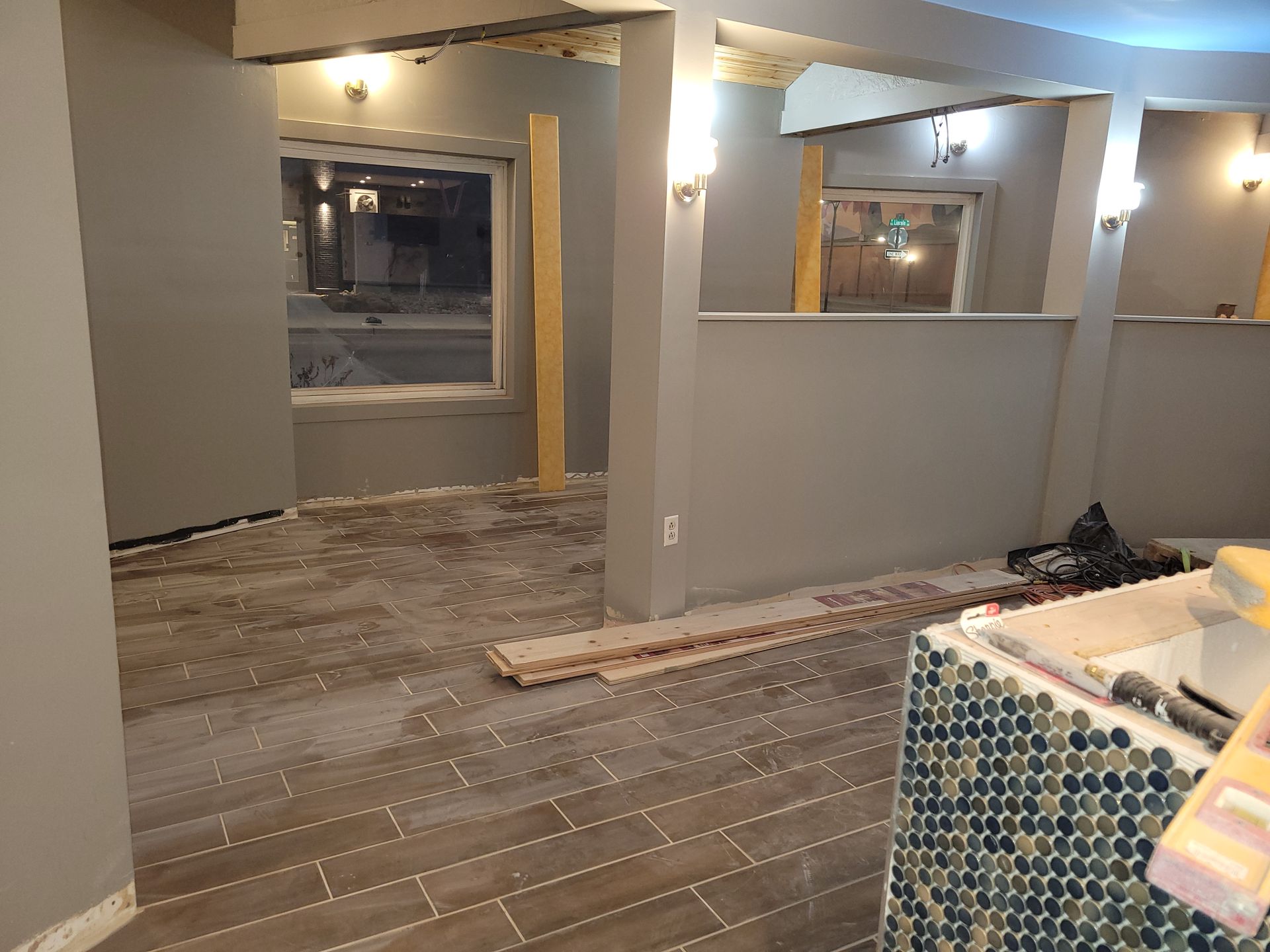 Partially renovated room with exposed subflooring, gray walls, and warm overhead lights, viewed toward windows