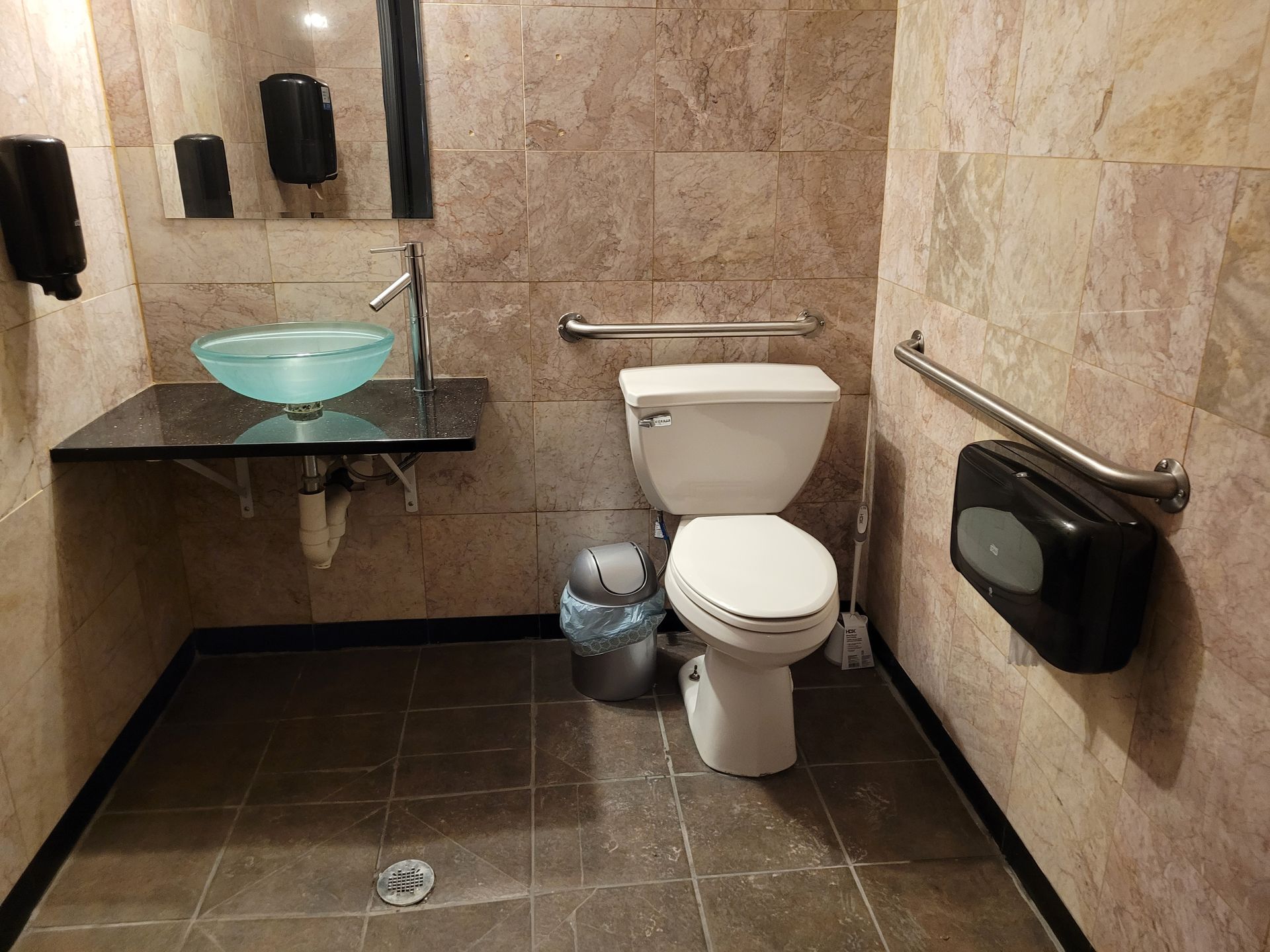 Small beige-tiled public restroom with a toilet, glass sink, trash can, and black soap dispensers.