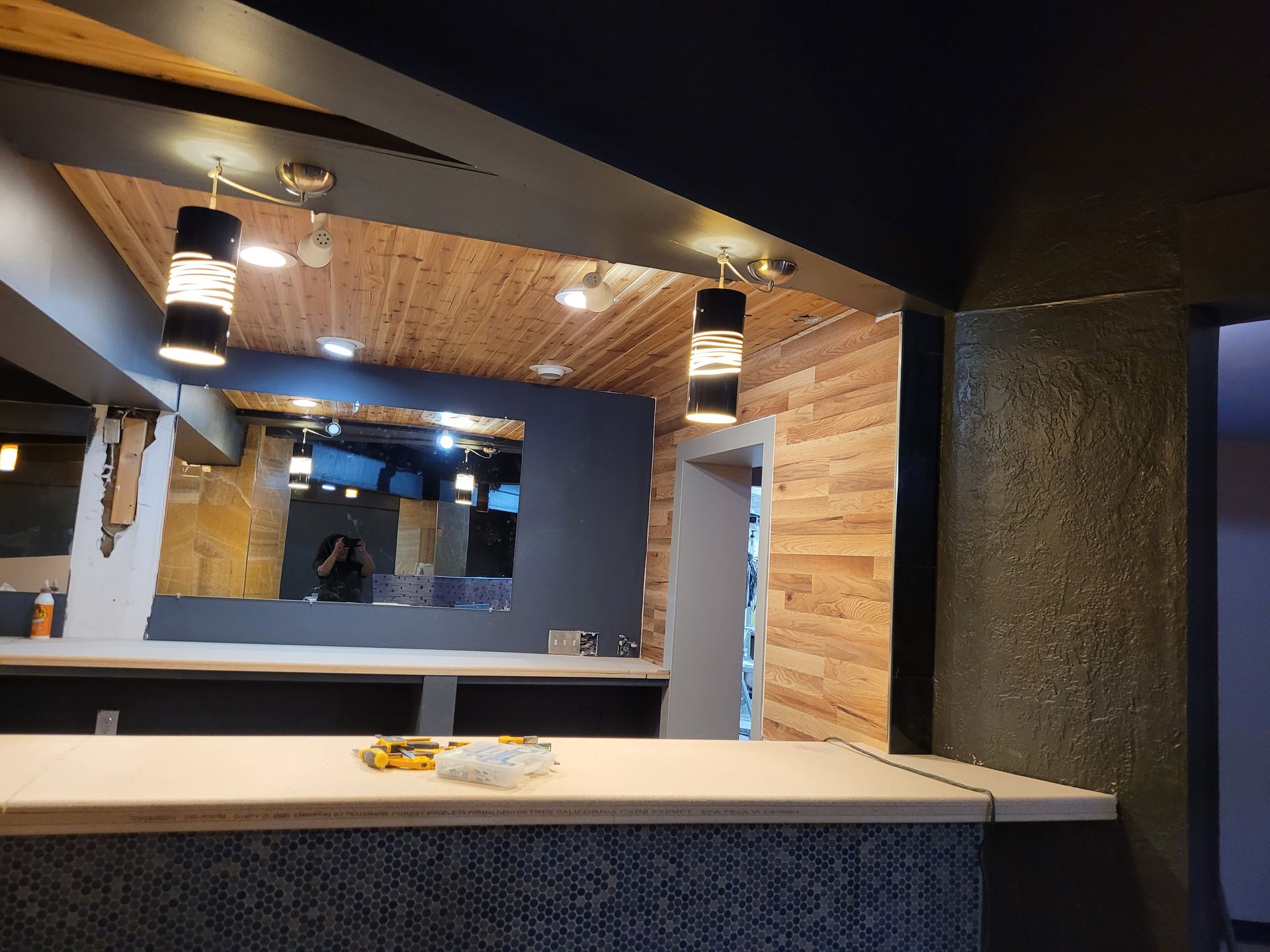 Modern bar interior with wood paneling, pendant lights, and a long counter beneath a large mirror