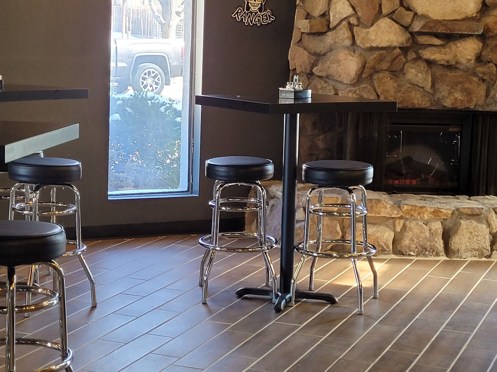 Rustic bar area with stools, wood floor, and stone fireplace by a bright window.