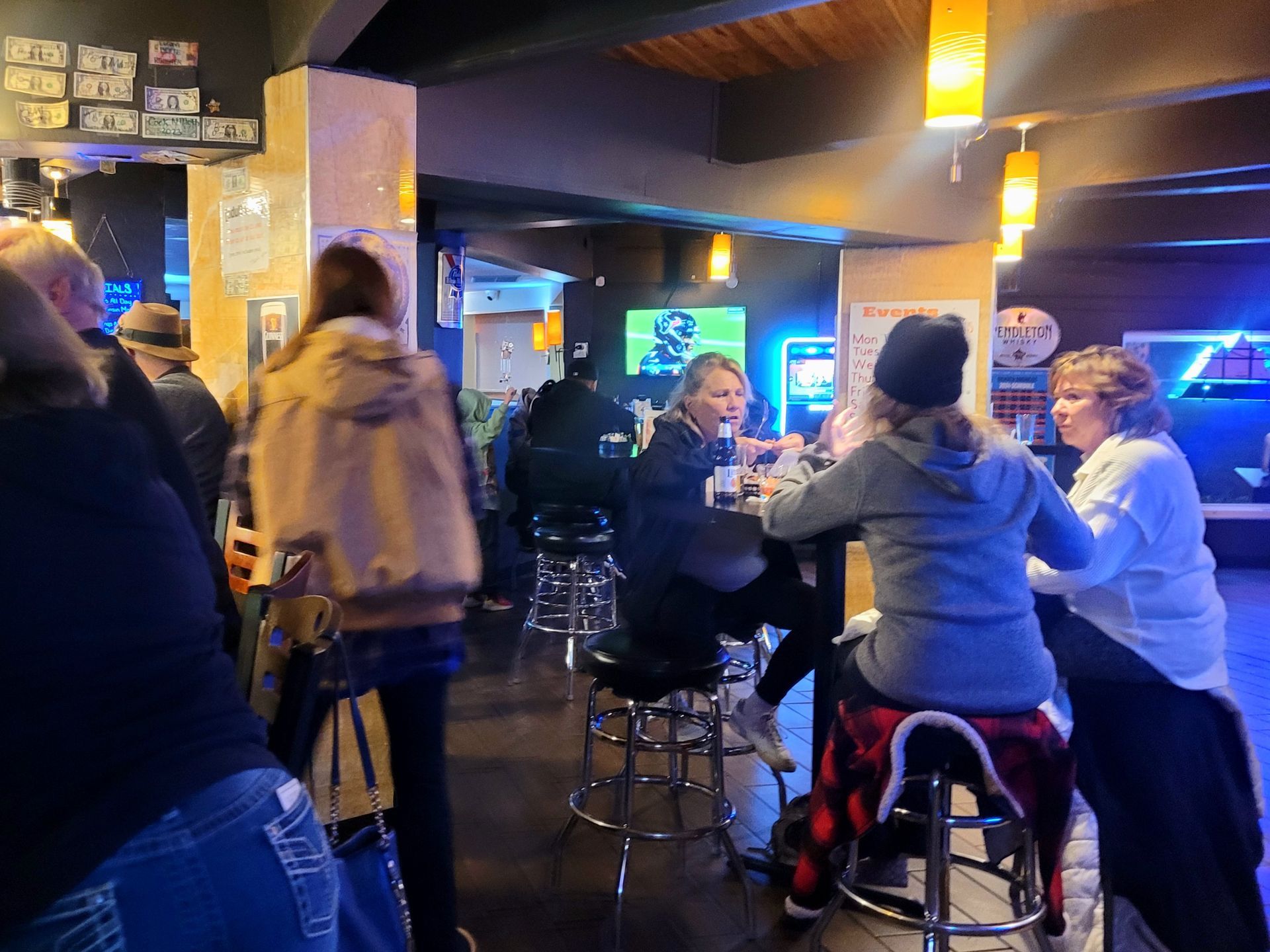 People chatting at a dimly lit bar with stools, glowing blue screens, and warm hanging lights